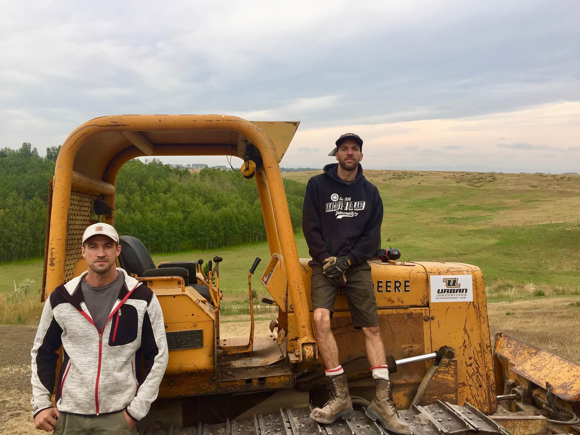 Two concrete workers pose for a picture with a vintage John Deere tractor on a rolling ridge of green grasses.