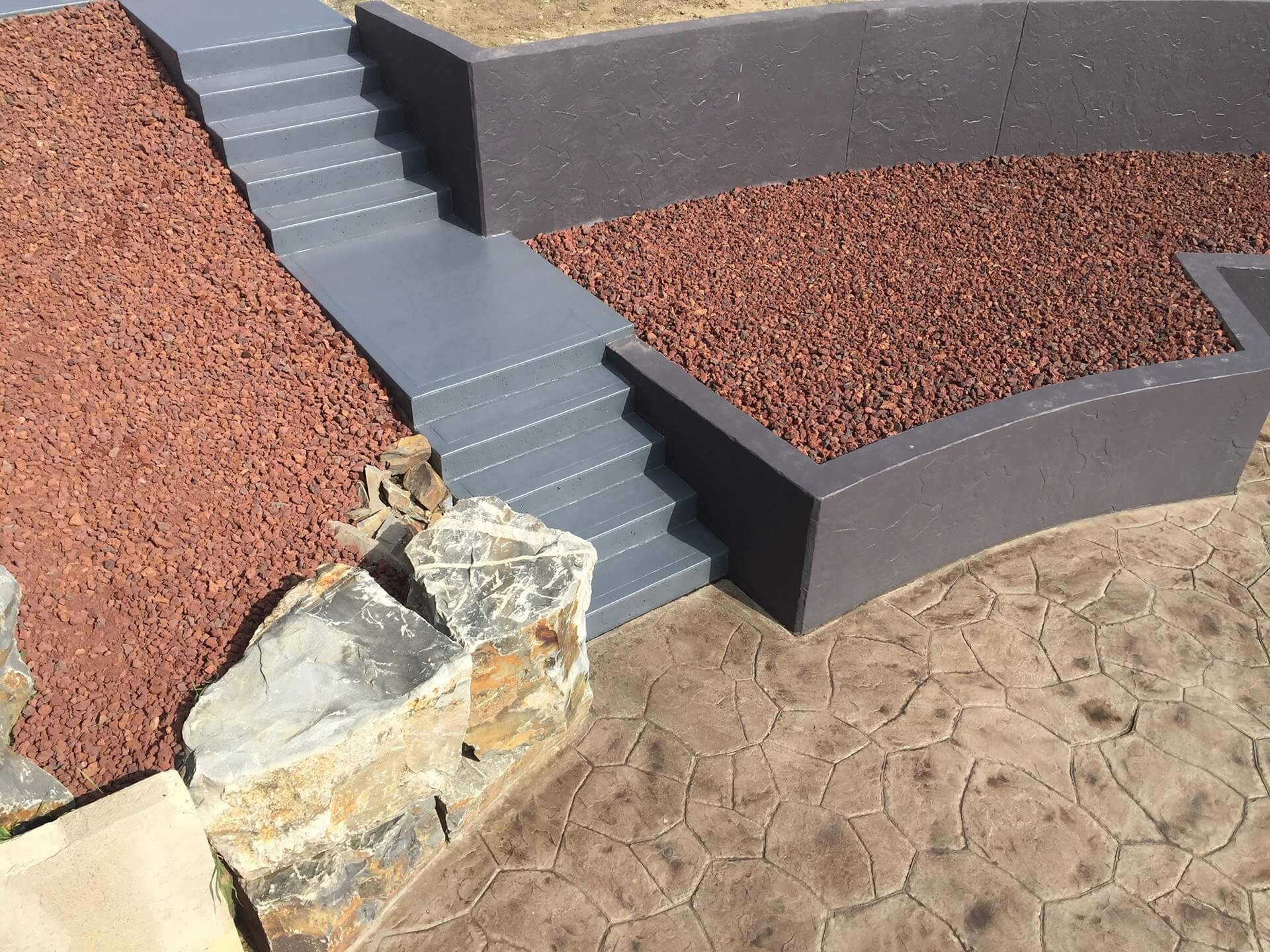 A concrete staircase leads down two retaining walls to a Sante Fe stamped concrete patio. Areas between the walls and patio are filled with red lava rock.