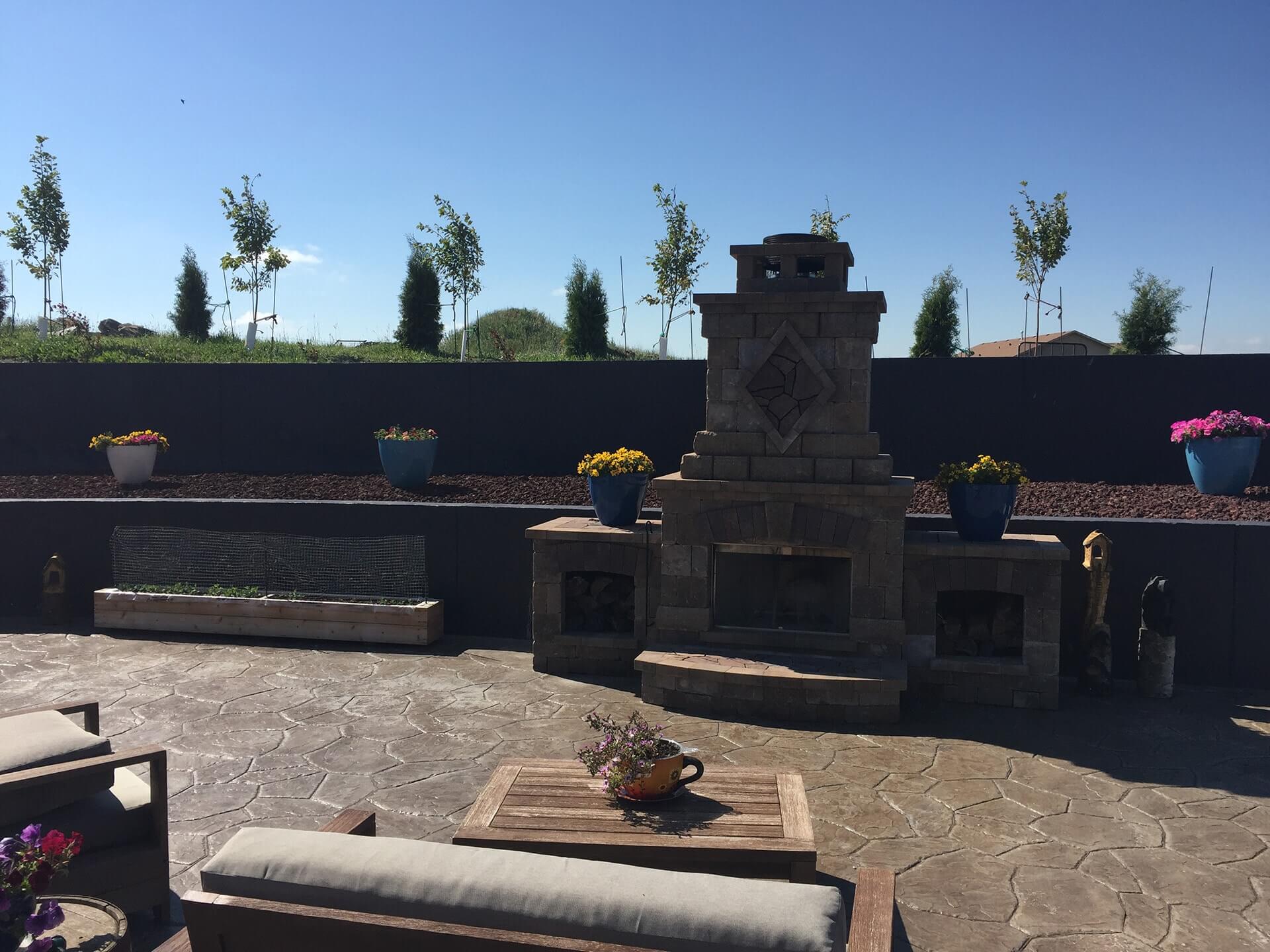 A large stone fireplace is surrounded with potted plants and patio furniture on a stamped concrete patio with retaining walls.