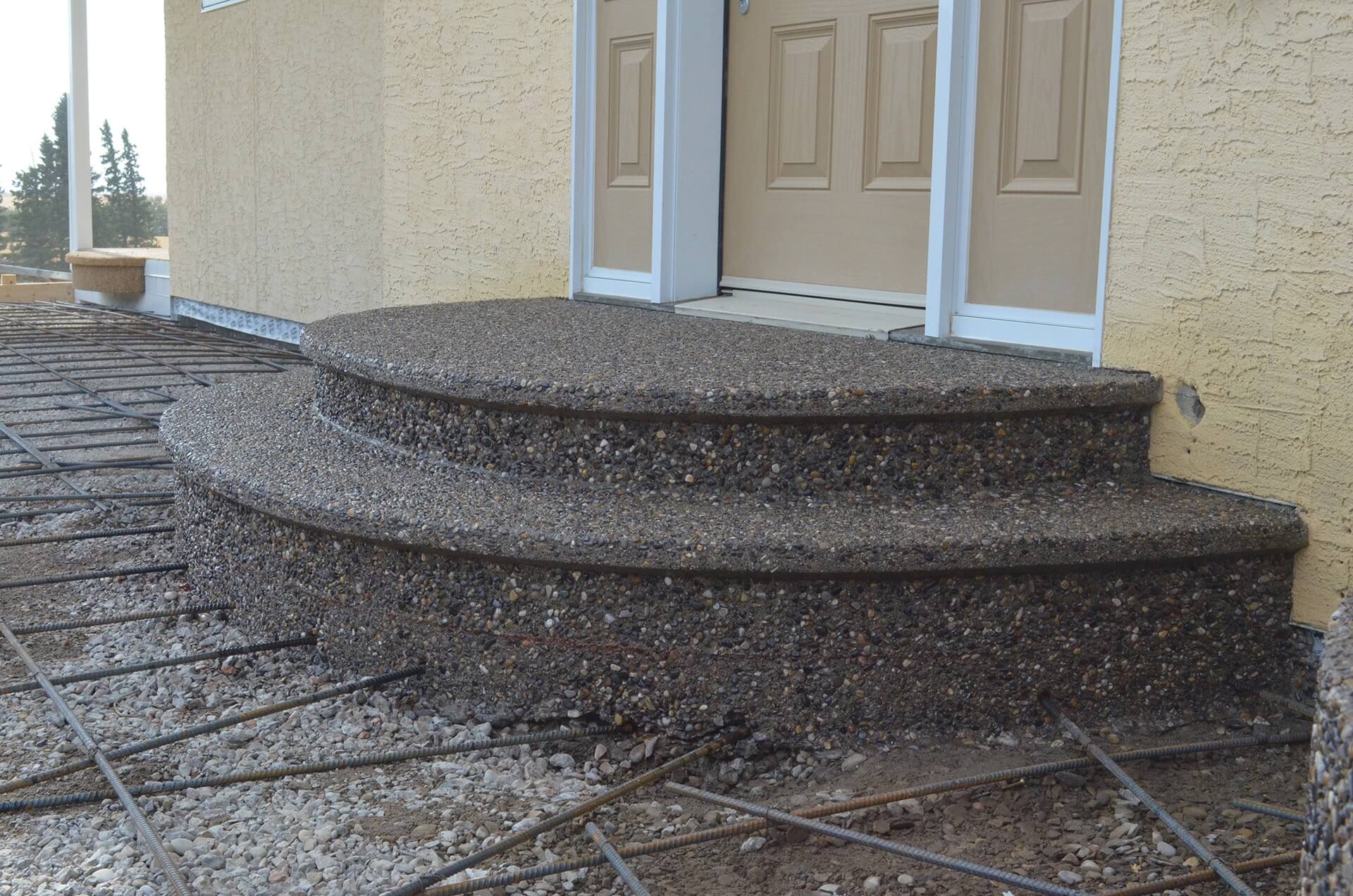 Exposed aggregate steps with curved faces and a bull nose are set at an entrance to a home. The surrounding rebar is set into them for reinforcement.