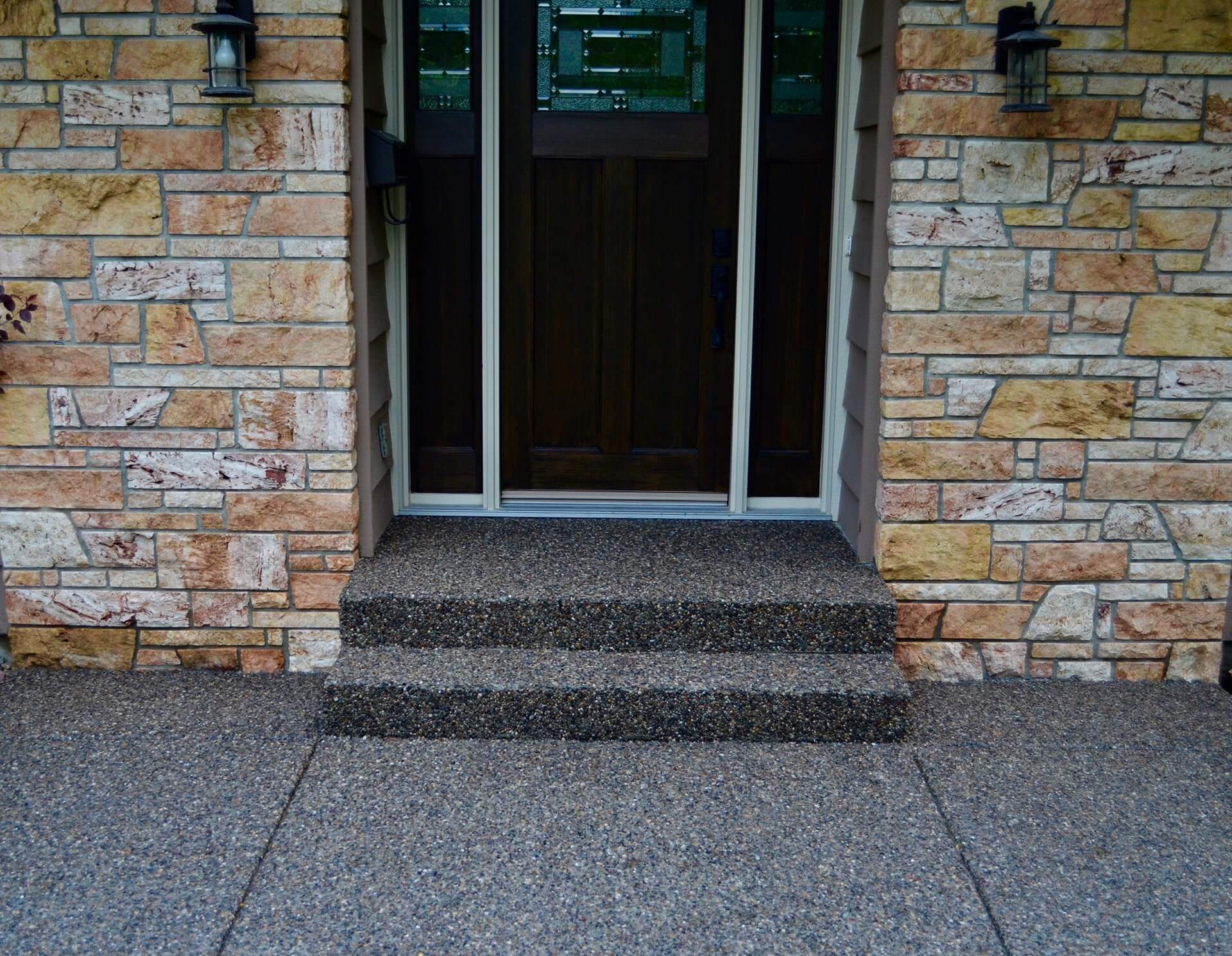 a two riser exposed aggregate front step and adjoining sidewalk frame the front entrance of a house with a black door and natural stone veneer.