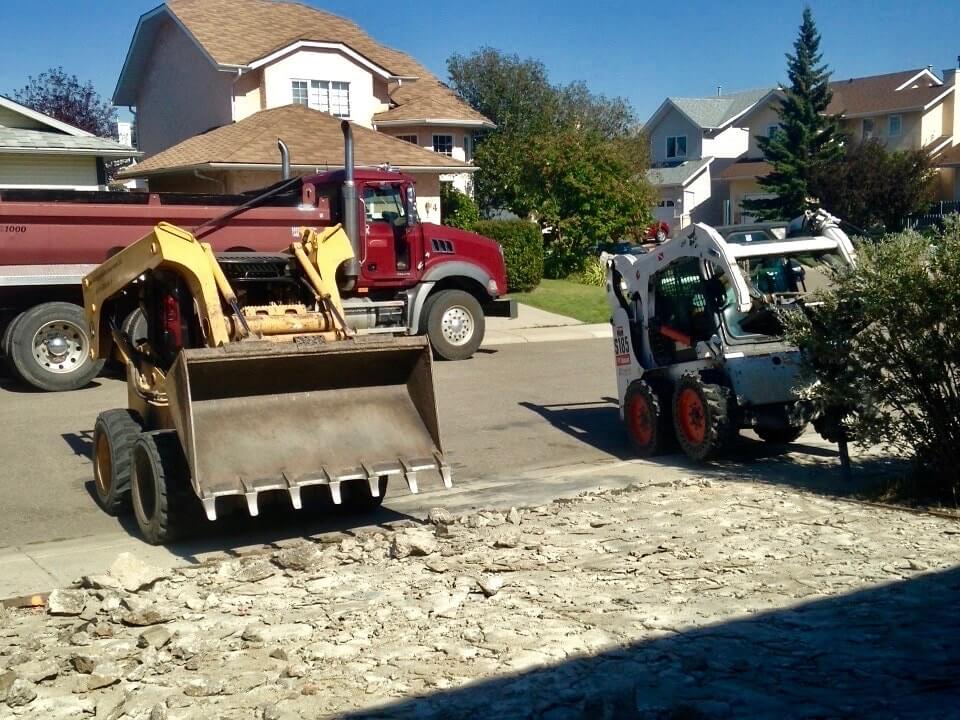 Two Bobcats work to hydraulically break and remove an old driveway. A dump truck waits on the street to load out the old concrete.
