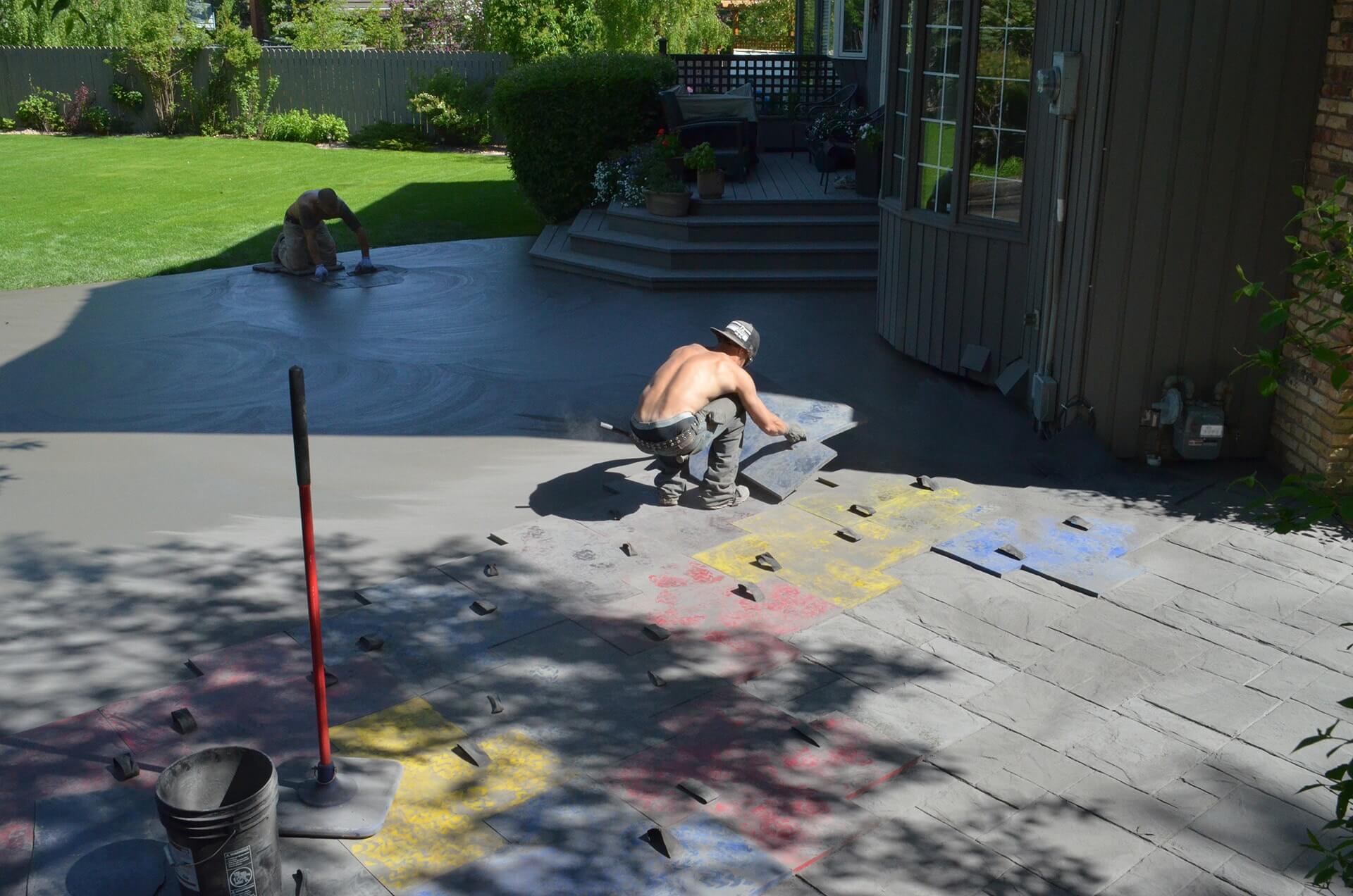 A concrete finisher works to place stamps in wet concrete. The impressions of the mats allow for patterns to achieved in concrete. Pigmented powder is visible and allows the mats not to stick to the fresh concrete while also allowing a stain to be absorbed for a realistic finish.