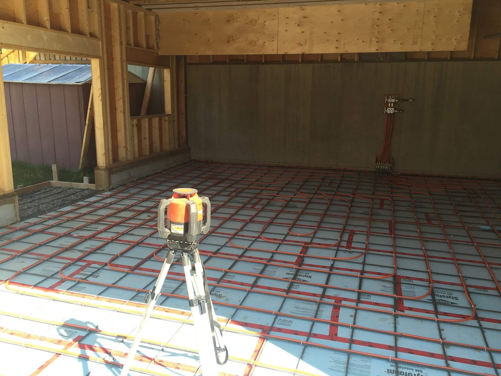 a rotating laser level sits atop a tripod in an engineered garage. The garage floor has insulation, rebar, and piping laid out for in slab heating.
